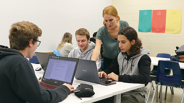 Une professeure donne une explication à des étudiants qui sont assis à leur bureau avec un portable.